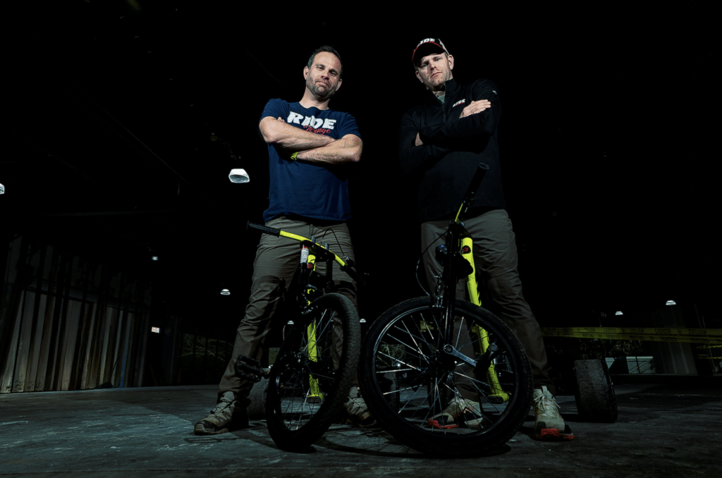 Two men standing with bikes in a darkened warehouse space with their arms crossed in front of them. From Ride Lounge Car Club.