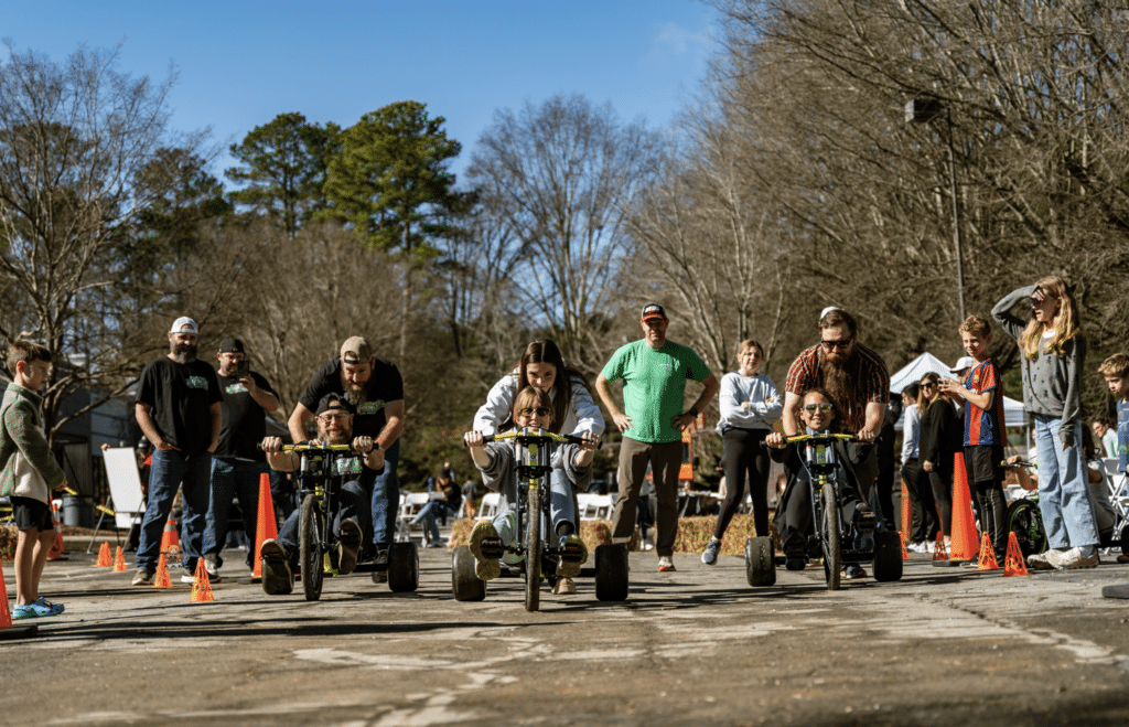 A group of kids with parents and spectators taking part in a trike race in a warehouse parking lot. Pedal Fest at Ride Lounge Car Club.