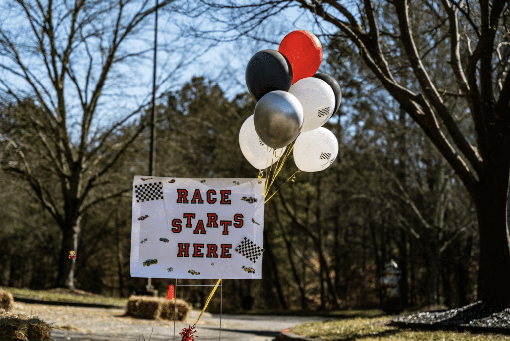 A handmade sign and white, red, black and grey balloons. The sign reads 'Race Starts Here' and is located in a warehouse parking lot with grass and trees in the background. From Ride Lounge car club.