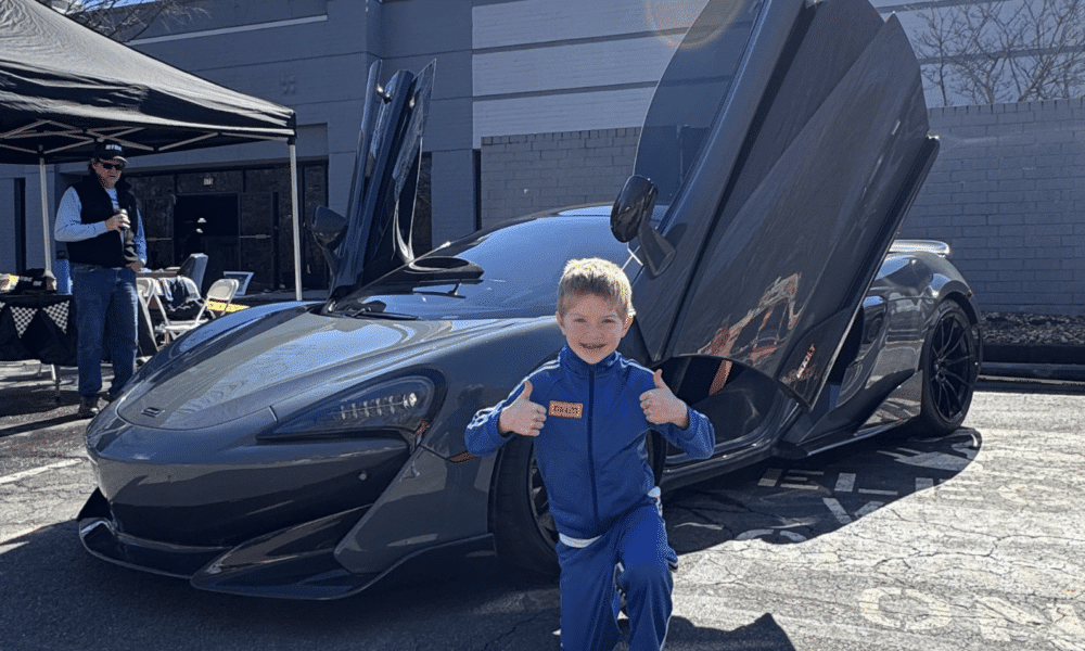 A smiling kid with blonde hair giving a thumbs up in front of an expensive sports car with it's driver door up.