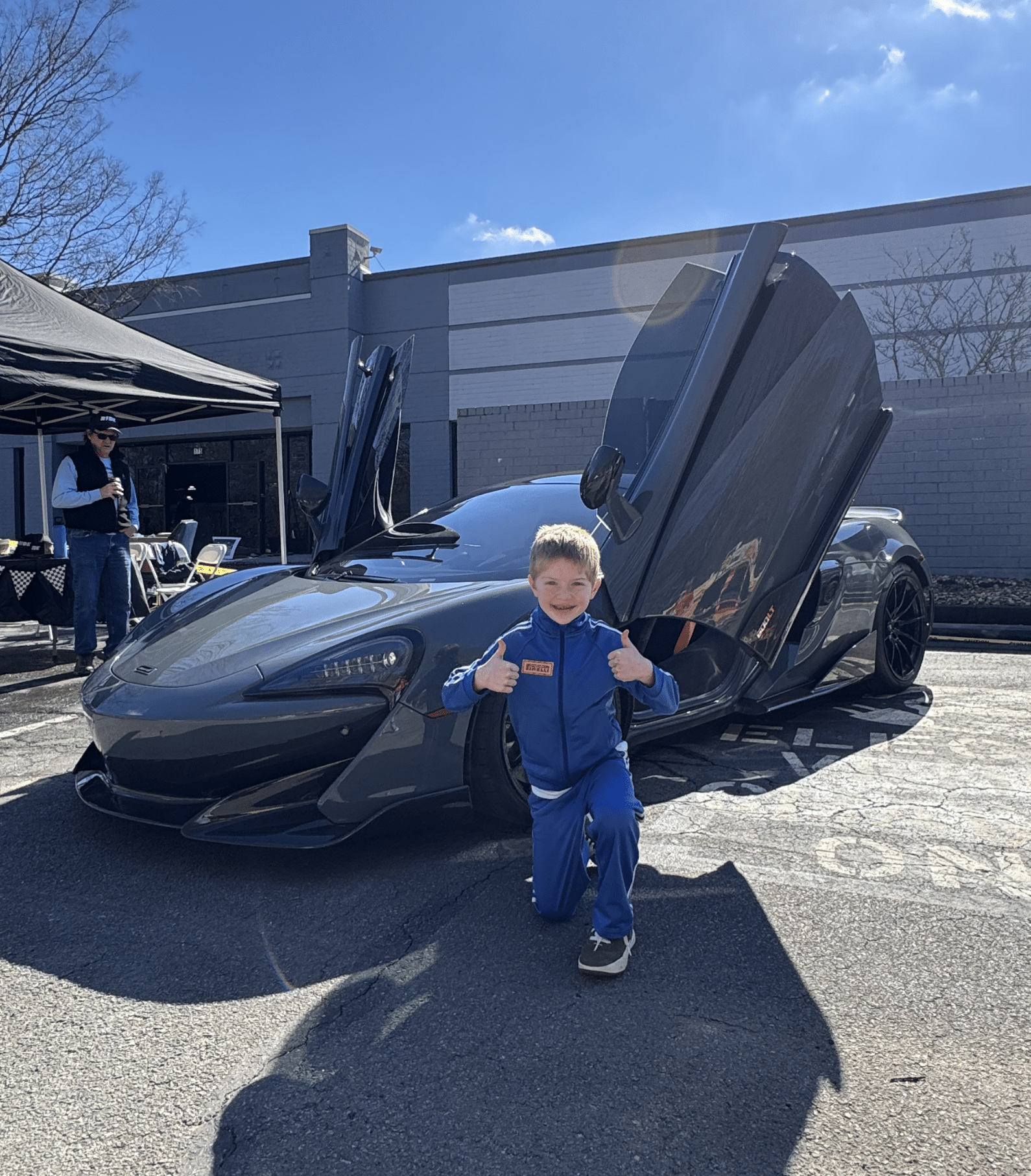 A smiling kid with blonde hair giving a thumbs up in front of an expensive sports car with it's driver door up.