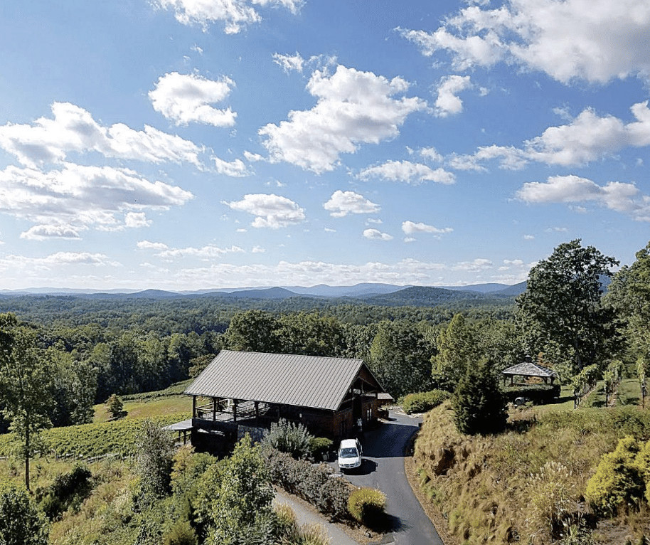 Expansive view of Wolf Winery from a hilltop. There's a building with parking surrounded by acres of trees and greenery. Blue sky dotted with fluffy white clouds overhead.