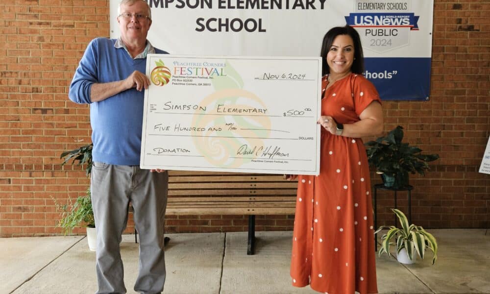 An older white man in a blue shirt holding a giant donation check with a young, dark-haired woman from Simpson Elementary School