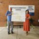 An older white man in a blue shirt holding a giant donation check with a young, dark-haired woman from Simpson Elementary School