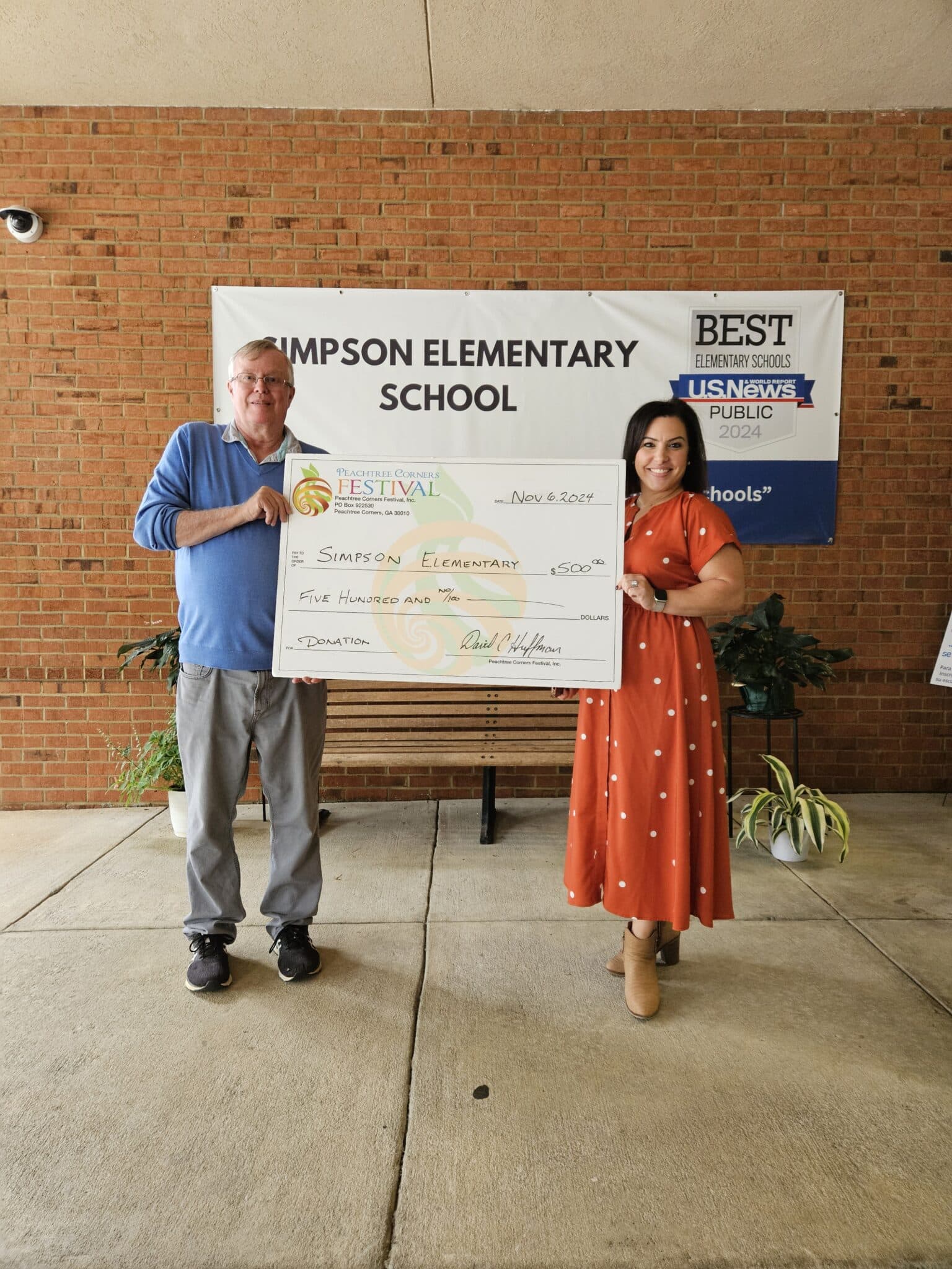 An older white man in a blue shirt holding a giant donation check with a young, dark-haired woman from Simpson Elementary School