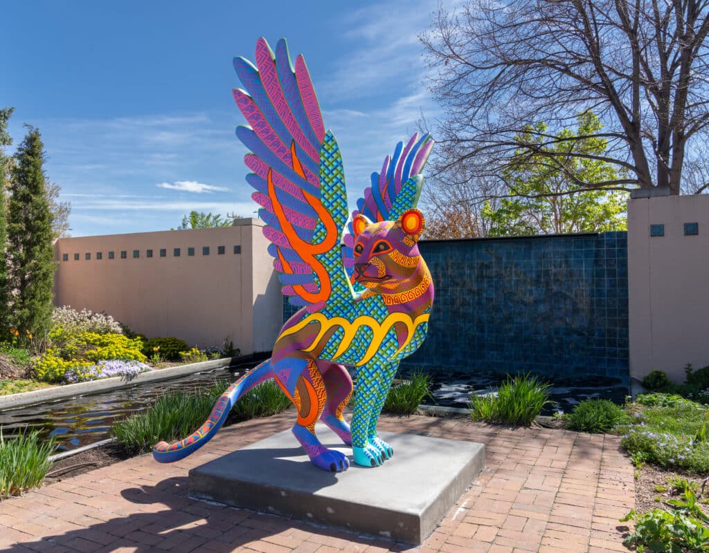 Large colorful sculpture of a winged mythical creature on an open patio in a wooded garden.