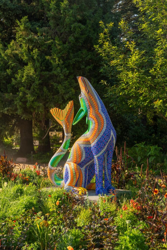 Large colorful sculpture of a howling fish-wolf mythical creature in a wooded garden.