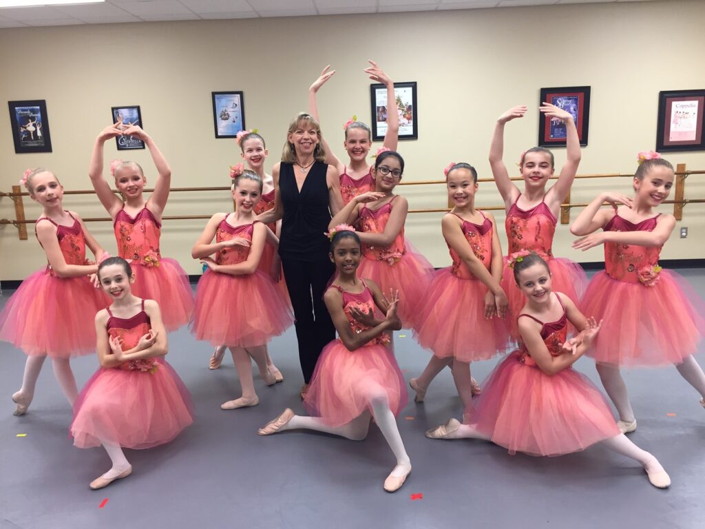 A middle aged woman with blonde hair, wearing all black, standing amidst a group of young ballerinas, posing and wearing pink tutus.