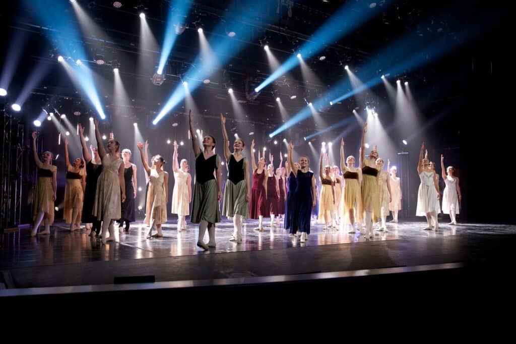 A group of dancers on stage for a finale. They all stand with an arm raised, looking up with blue and white lighting.