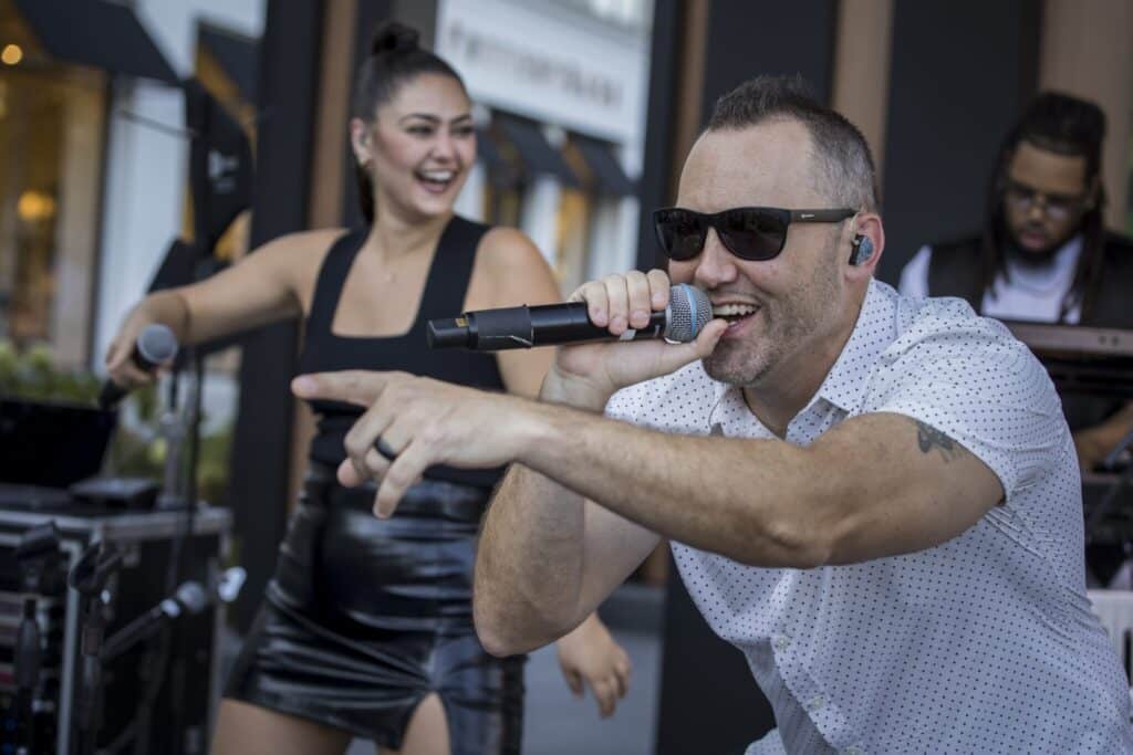 A band performing on an outdoor stage. A man with short hair, white t-shirt and sunglasses sings into the mic and points to the crowd. A woman with dark hair and short, black dress is in the background smiling. And other musicians are behind them.