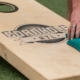 Close-up of a cornhole board and a hand reaching down to pick up blue/green beanbags.