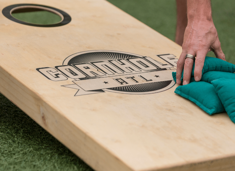 Close-up of a cornhole board and a hand reaching down to pick up blue/green beanbags.