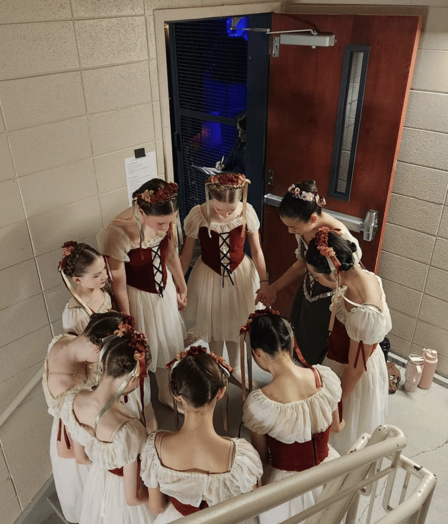 a group of young ballet dancers standing in a stairwell next to an open door. They are in a circle with heads bowed, praying before going onstage for their performance.