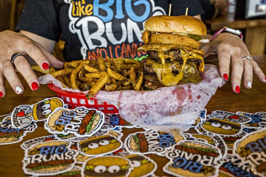 Woman with red and white painted nails sitting at a table covered with Burger Week stickers. There's a basket in front of her that has a large cheese burger and fries.