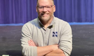 A middle-aged man with short hair and glasses. He's wearing a grey long-sleeved shirt and his arms are crossed in front of him. He's standing in front of an empty high school theatre stage with a purple curtain behind him.