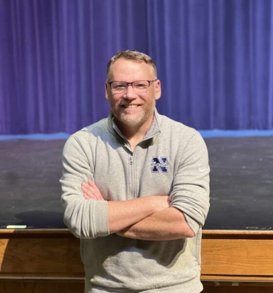 A middle-aged man with short hair and glasses. He's wearing a grey long-sleeved shirt and his arms are crossed in front of him. He's standing in front of an empty high school theatre stage with a purple curtain behind him.