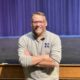 A middle-aged man with short hair and glasses. He's wearing a grey long-sleeved shirt and his arms are crossed in front of him. He's standing in front of an empty high school theatre stage with a purple curtain behind him.