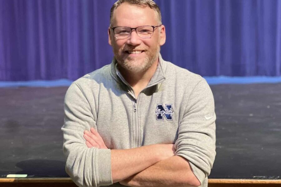 A middle-aged man with short hair and glasses. He's wearing a grey long-sleeved shirt and his arms are crossed in front of him. He's standing in front of an empty high school theatre stage with a purple curtain behind him.