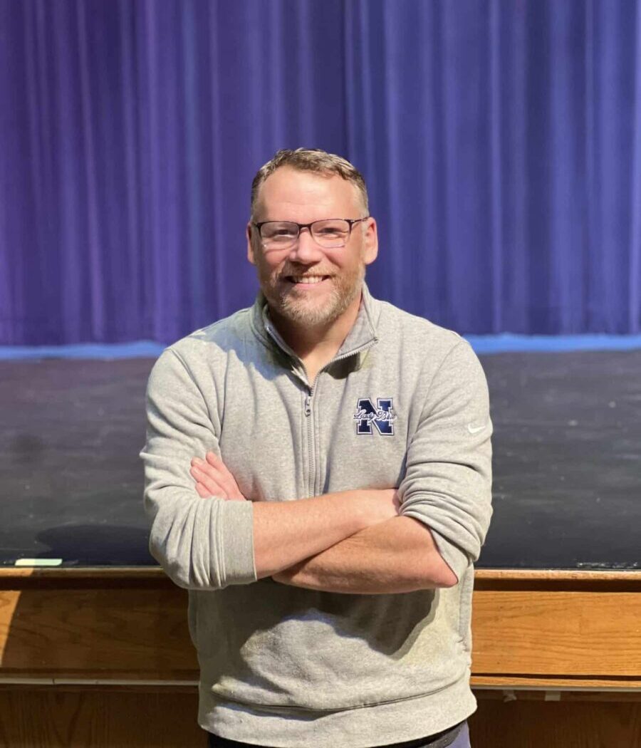 A middle-aged man with short hair and glasses. He's wearing a grey long-sleeved shirt and his arms are crossed in front of him. He's standing in front of an empty high school theatre stage with a purple curtain behind him.