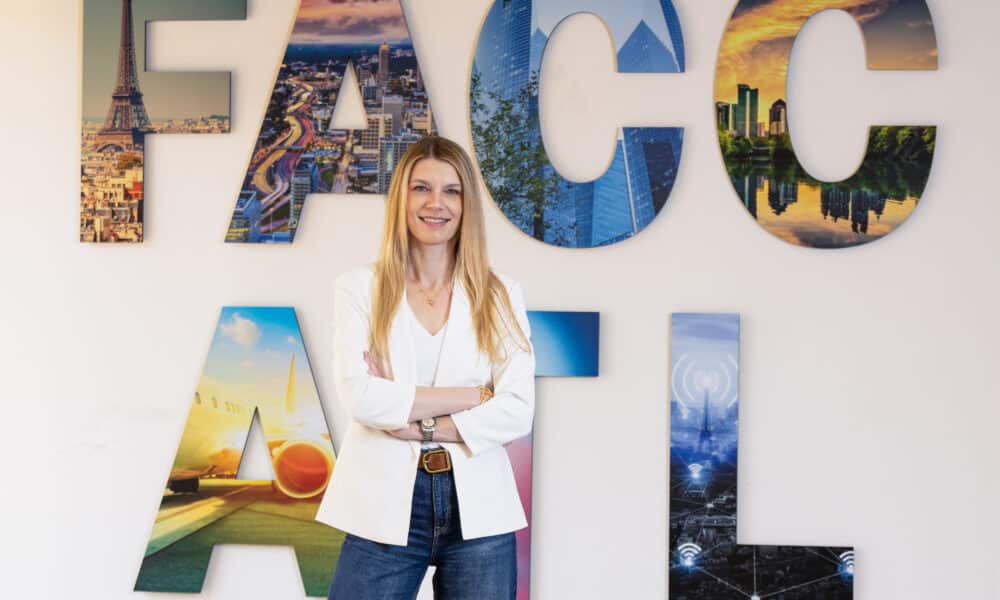 A woman with long blonde hair, wearing a white top and white jacket with blue jeans. She's standing in front of a wall with FACC ATL in large letters.