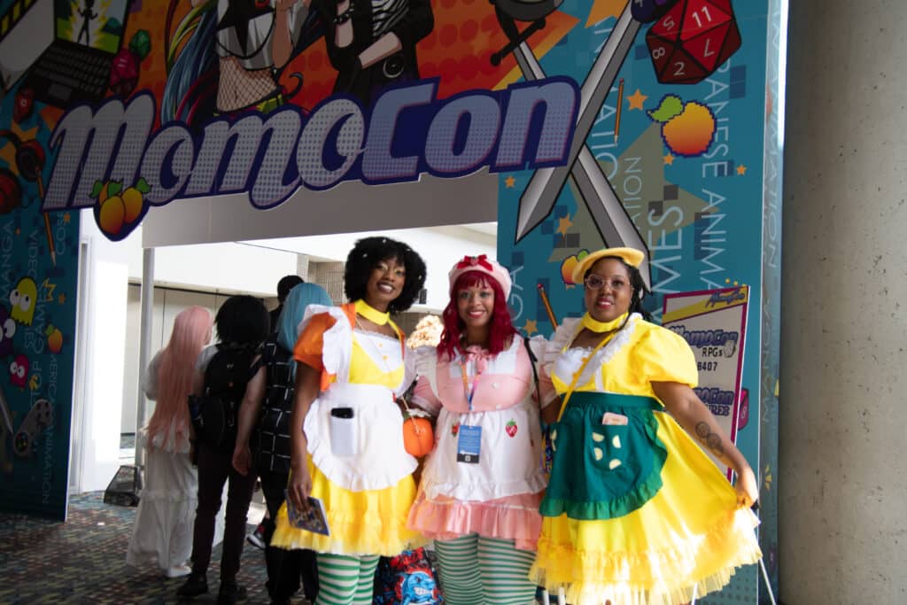 Three young Black women wearing cosplay and standing at the entrance of MomoCon 2025