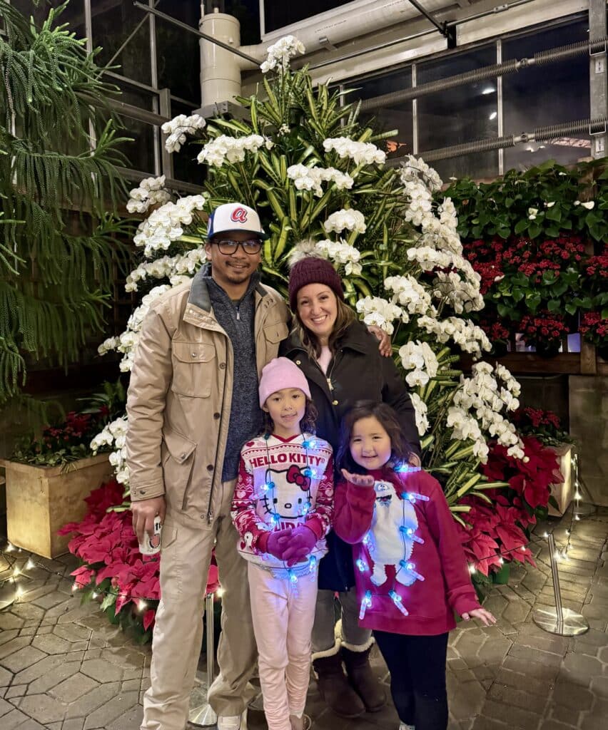 A mom, dad and two young daughters standing in front of a display of holiday trees and plants.
