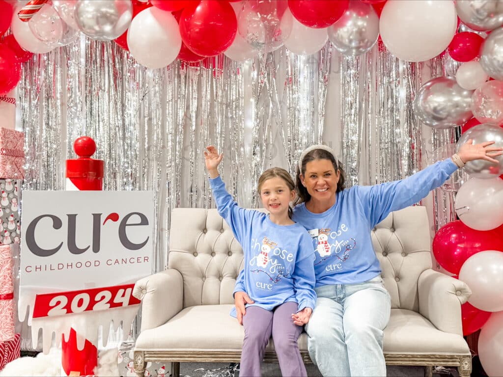 A mom and young daughter, both with dark hair and wearing blue shirts. They are sitting on a white sofa with streamers and red and white balloons in the background. There is a sign promoting Cure Childhood Cancer 2024.