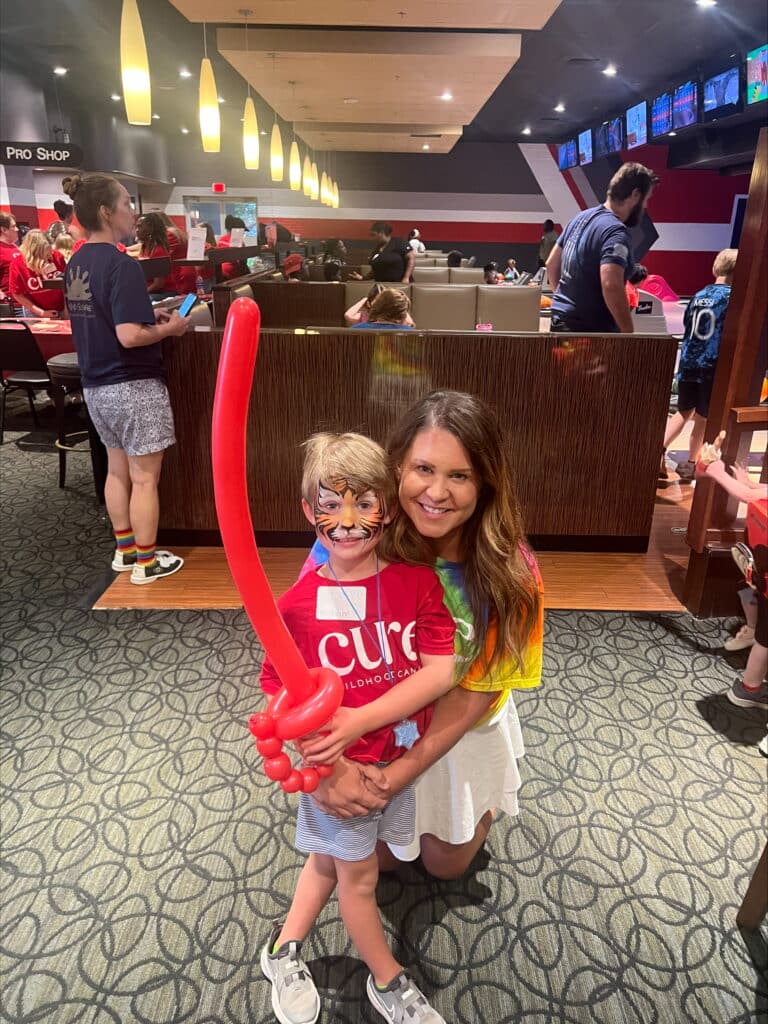 A mom with long dark hair crouched down behind her young son, who is wearing face paint and a red shirt. She's giving him a hug as they pose for a photo. He's holding a red balloon sword.
