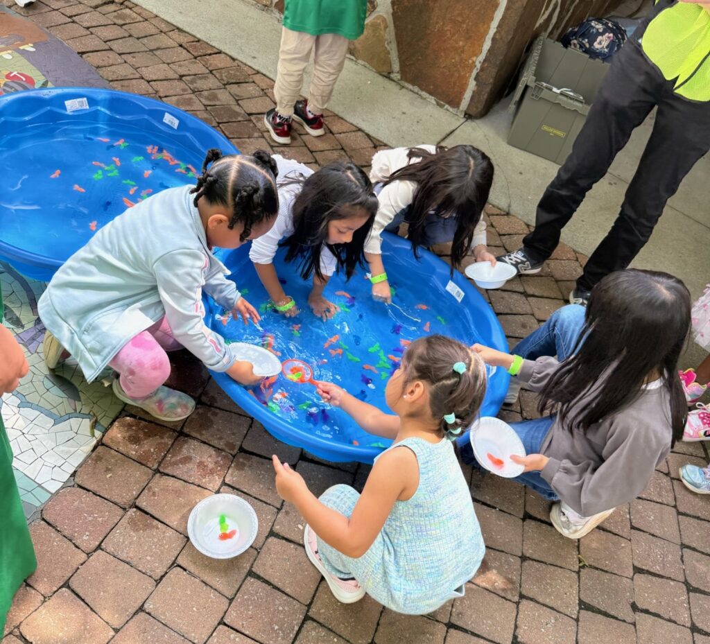 A group of children sitting on a bricked pathway in a park, playing a game at a Japan's Children's Day Festival in Norcross, Georgia