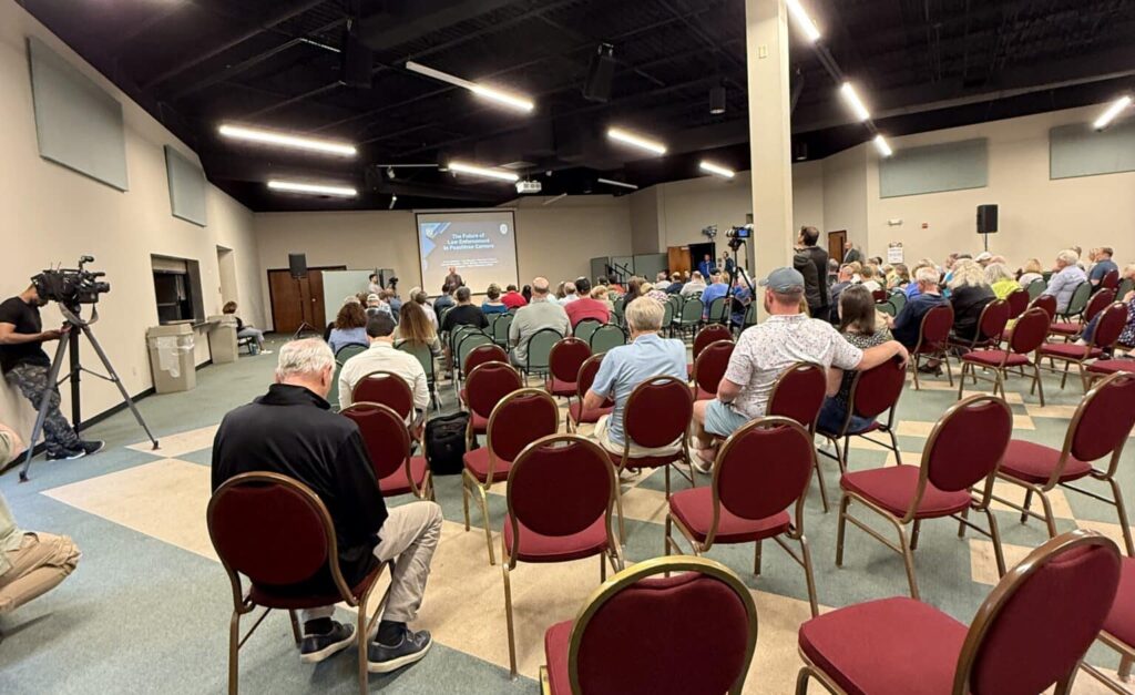 Audience and speaker, along with a few empty red chairs, at a city meeting discussing starting a police department