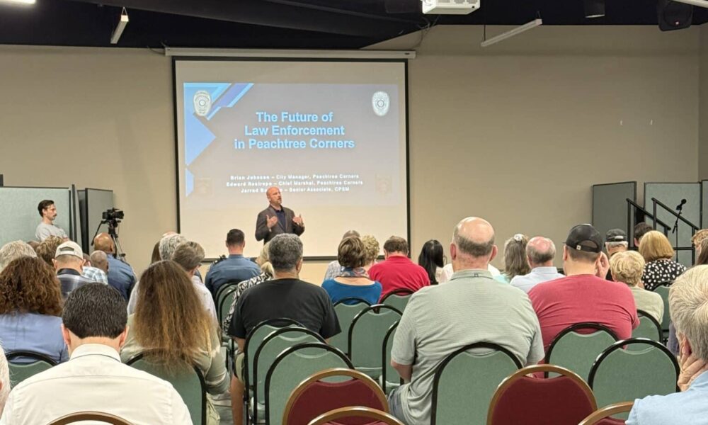 A group of people in the audience of a city meeting. They are sitting on red chairs, listening to a middle-aged white man at the front discussing the pros and cons of starting a police department in the city.