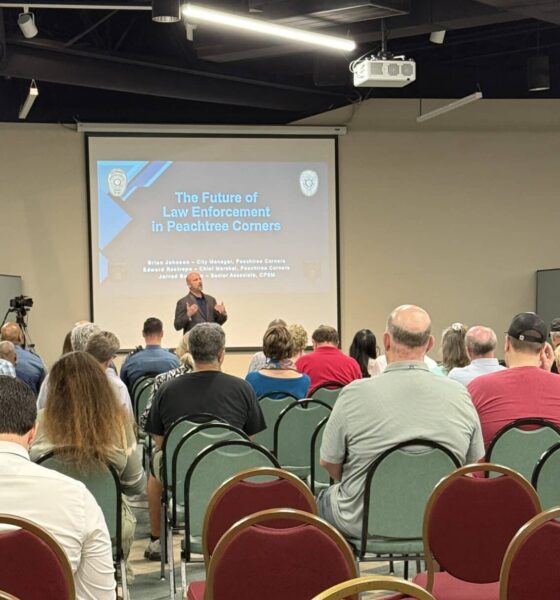 A group of people in the audience of a city meeting. They are sitting on red chairs, listening to a middle-aged white man at the front discussing the pros and cons of starting a police department in the city.