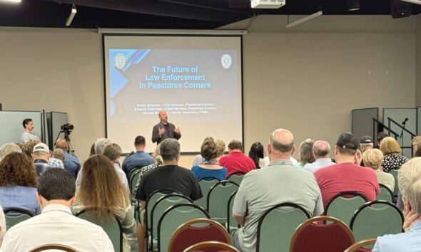 A group of people in the audience of a city meeting. They are sitting on red chairs, listening to a middle-aged white man at the front discussing the pros and cons of starting a police department in the city.