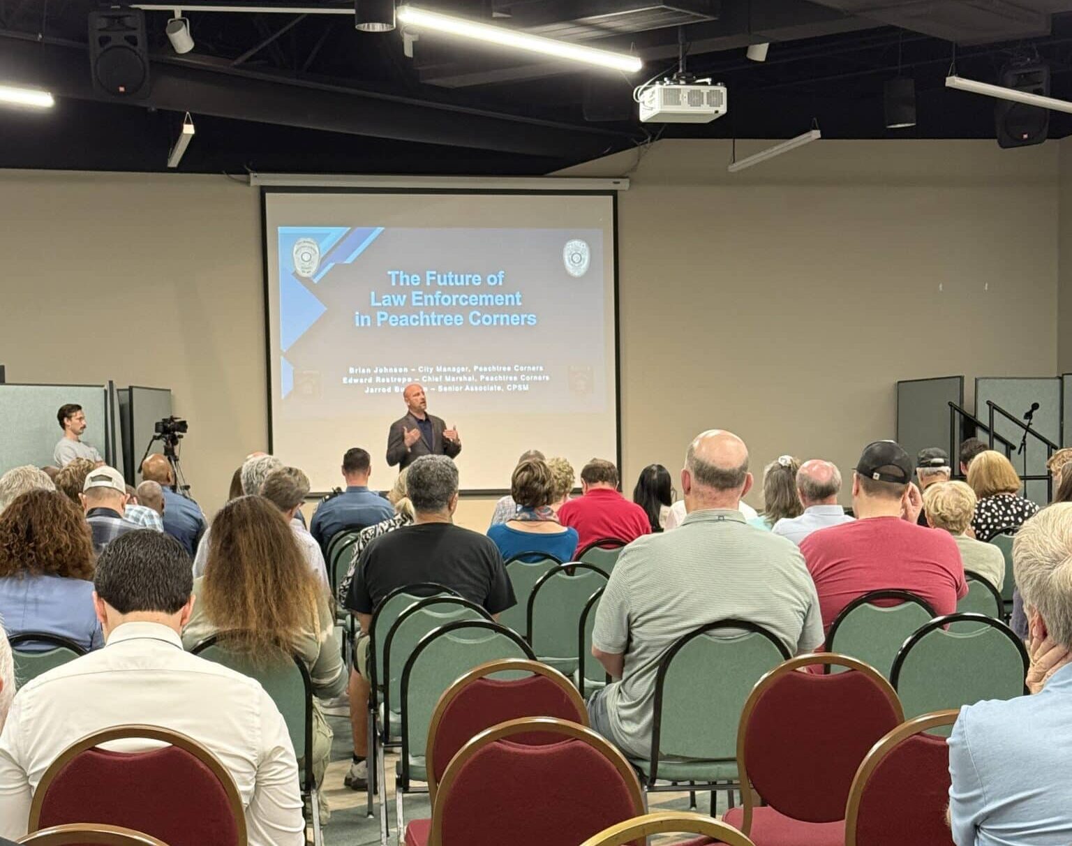 A group of people in the audience of a city meeting. They are sitting on red chairs, listening to a middle-aged white man at the front discussing the pros and cons of starting a police department in the city.