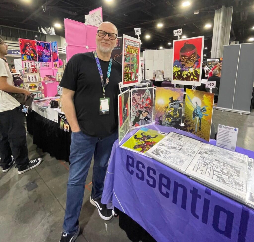 Middle aged man wearing black tshirt and jeans standing in front of his comic creator booth in the artist alley at MomoCon 2025.