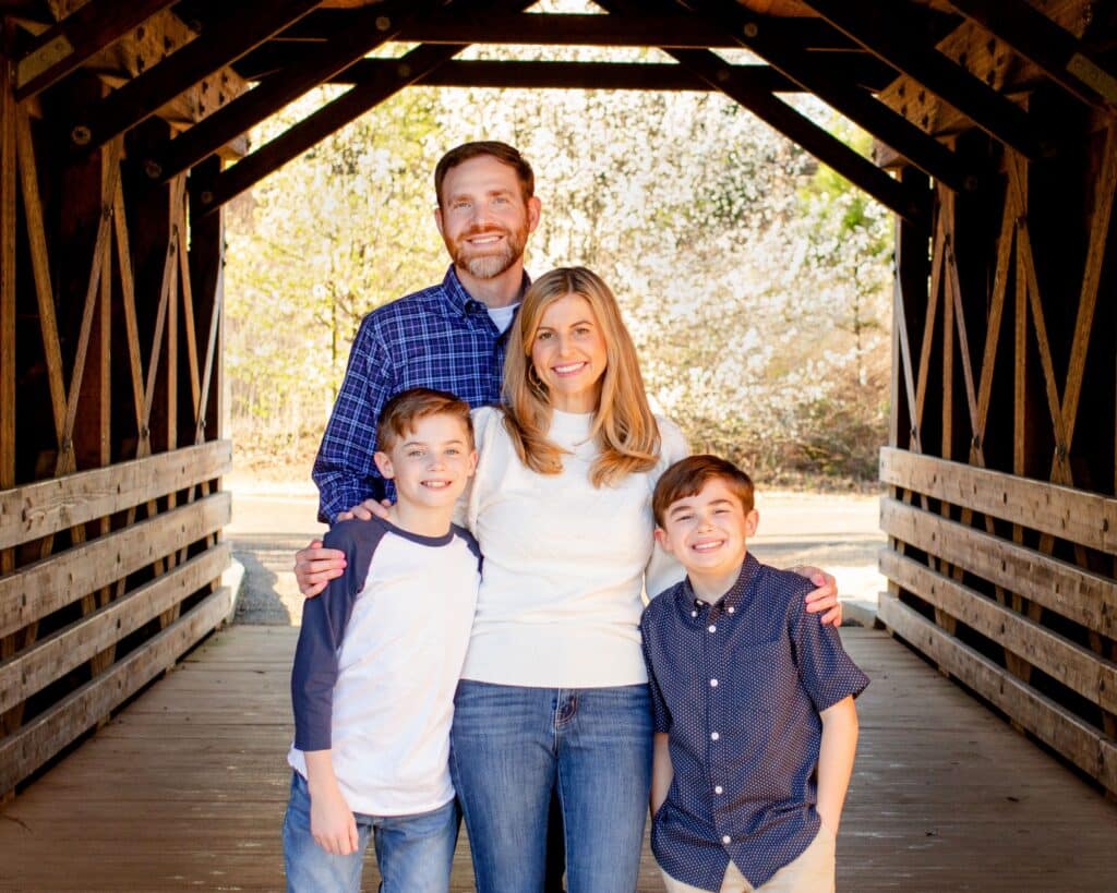 A mom with blonde hair and white shirt standing on a covered bridge with her husband who is wearing a dark blue shirt and her two young sons. They are all smiling.