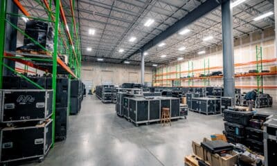 Interior of a spacious, clean and well-organized music production warehouse. There are tall ceilings with rows of lights, metal shelves full of audio/visual equipment and larger pieces of equipment grouped on the floor throughout the space.