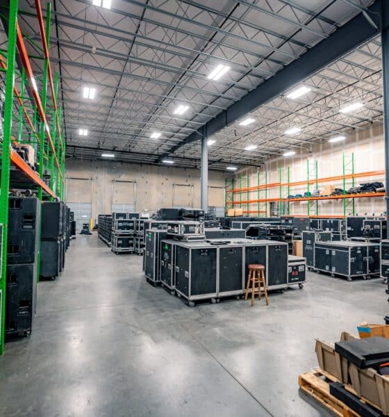 Interior of a spacious, clean and well-organized music production warehouse. There are tall ceilings with rows of lights, metal shelves full of audio/visual equipment and larger pieces of equipment grouped on the floor throughout the space.