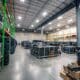 Interior of a spacious, clean and well-organized music production warehouse. There are tall ceilings with rows of lights, metal shelves full of audio/visual equipment and larger pieces of equipment grouped on the floor throughout the space.