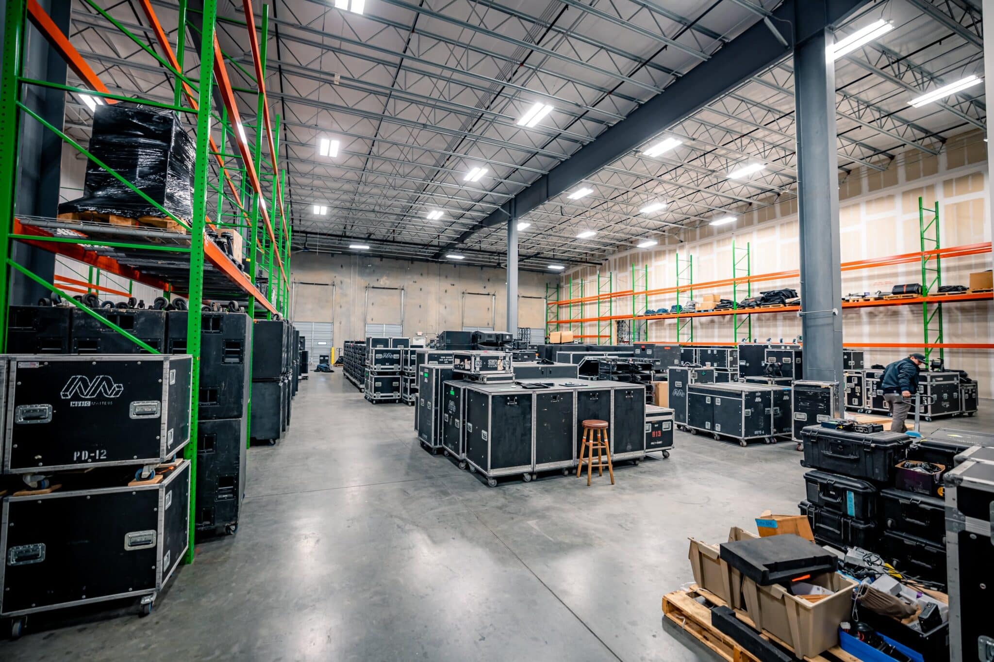 Interior of a spacious, clean and well-organized music production warehouse. There are tall ceilings with rows of lights, metal shelves full of audio/visual equipment and larger pieces of equipment grouped on the floor throughout the space.
