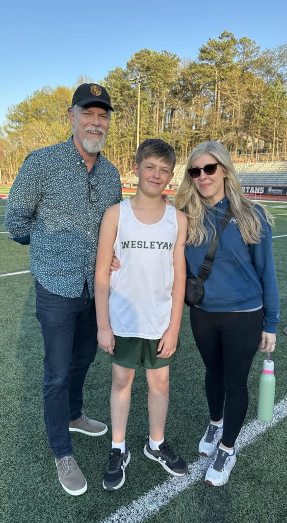 A man and woman standing outdoors on an athletic field with their pre-teen son. All are dressed casually. The son is wearing a white tank top and green shorts.