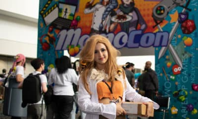 A young woman dressed in anime cosplay standing at the entrance to MomoCon 2024. The colorful convention logo is displayed over the entrance archway.