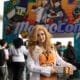 A young woman dressed in anime cosplay standing at the entrance to MomoCon 2024. The colorful convention logo is displayed over the entrance archway.