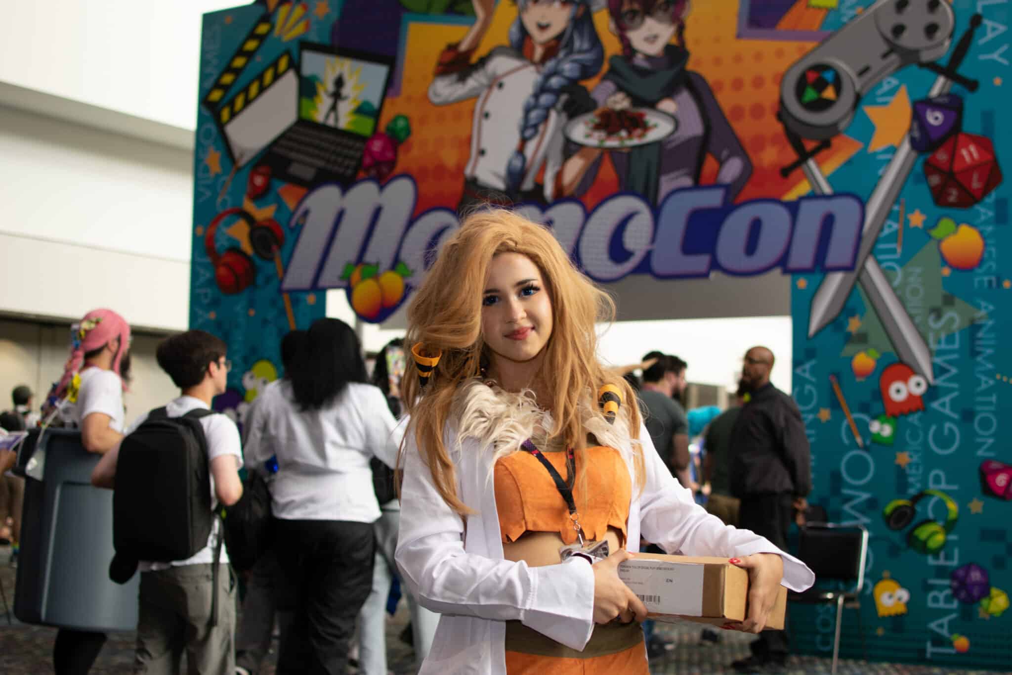 A young woman dressed in anime cosplay standing at the entrance to MomoCon 2024. The colorful convention logo is displayed over the entrance archway.