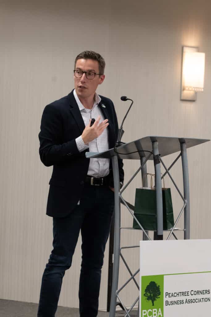 A man with short dark hair and glasses on stage behind a podium talking to the audience at a business event