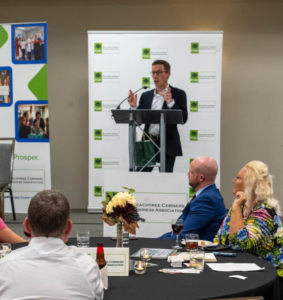 Bertrand Lapoire of Peachtree Corners standing on stage at a podium speaking to the audience at a PCBA business event.