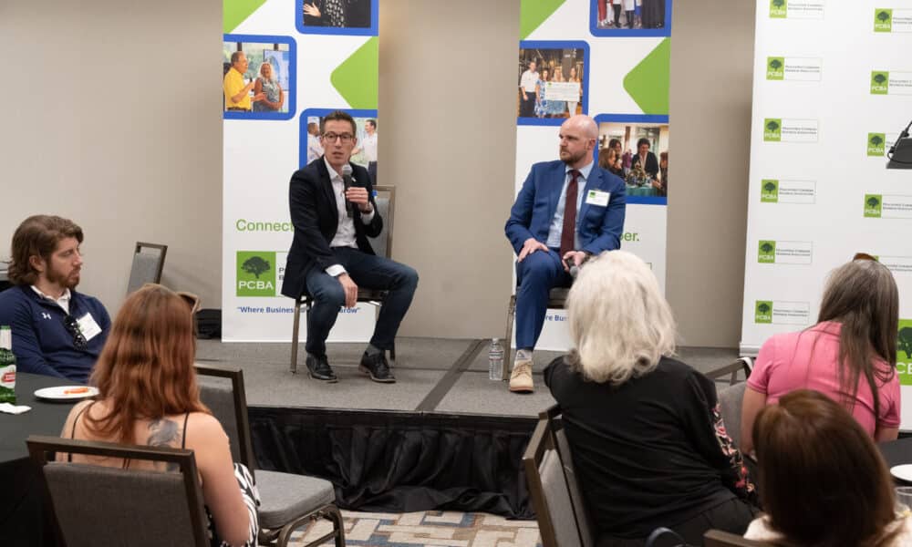 Two men sitting on chairs on a stage. They are holding microphones and answering questions from the audience at a business event