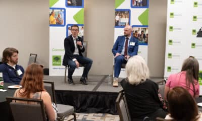 Two men sitting on chairs on a stage. They are holding microphones and answering questions from the audience at a business event