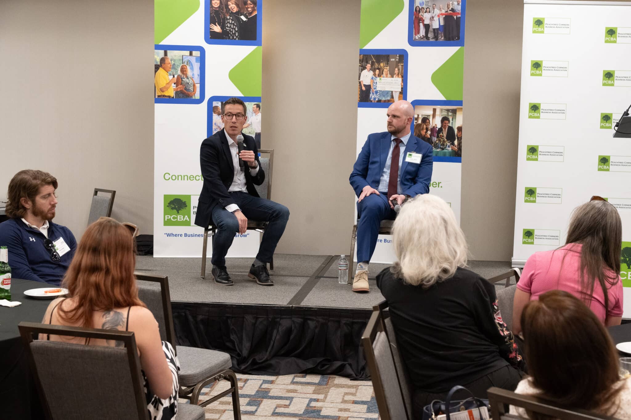 Two men sitting on chairs on a stage. They are holding microphones and answering questions from the audience at a business event