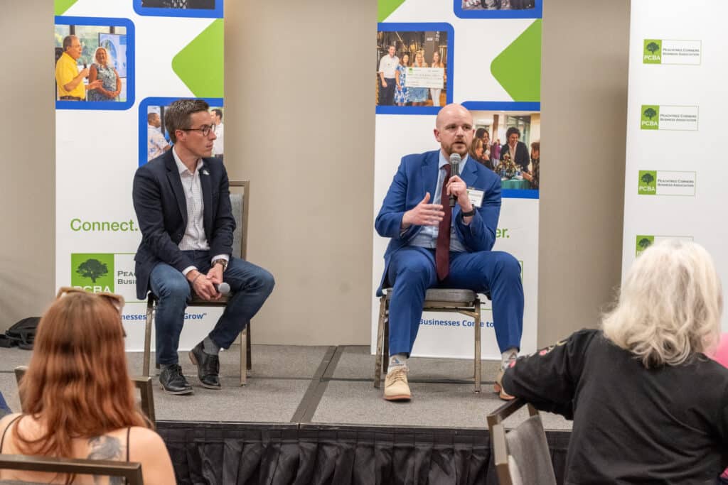 Two men sitting in chairs on stage at a business event. They are holding microphones and speaking to the audience